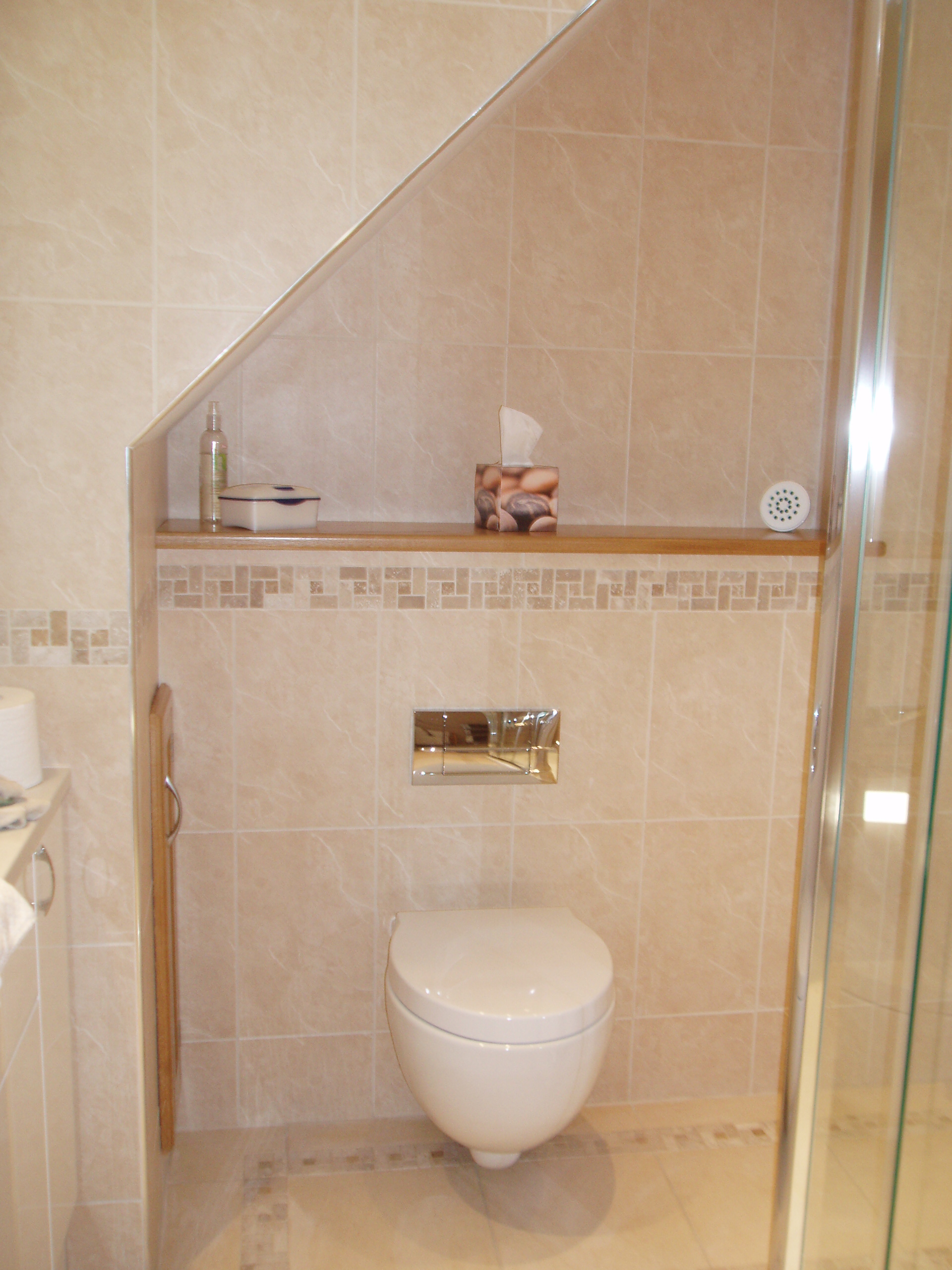 Wall-hung toilet beneath a sloping ceiling with a tiled shelf and mosaic border