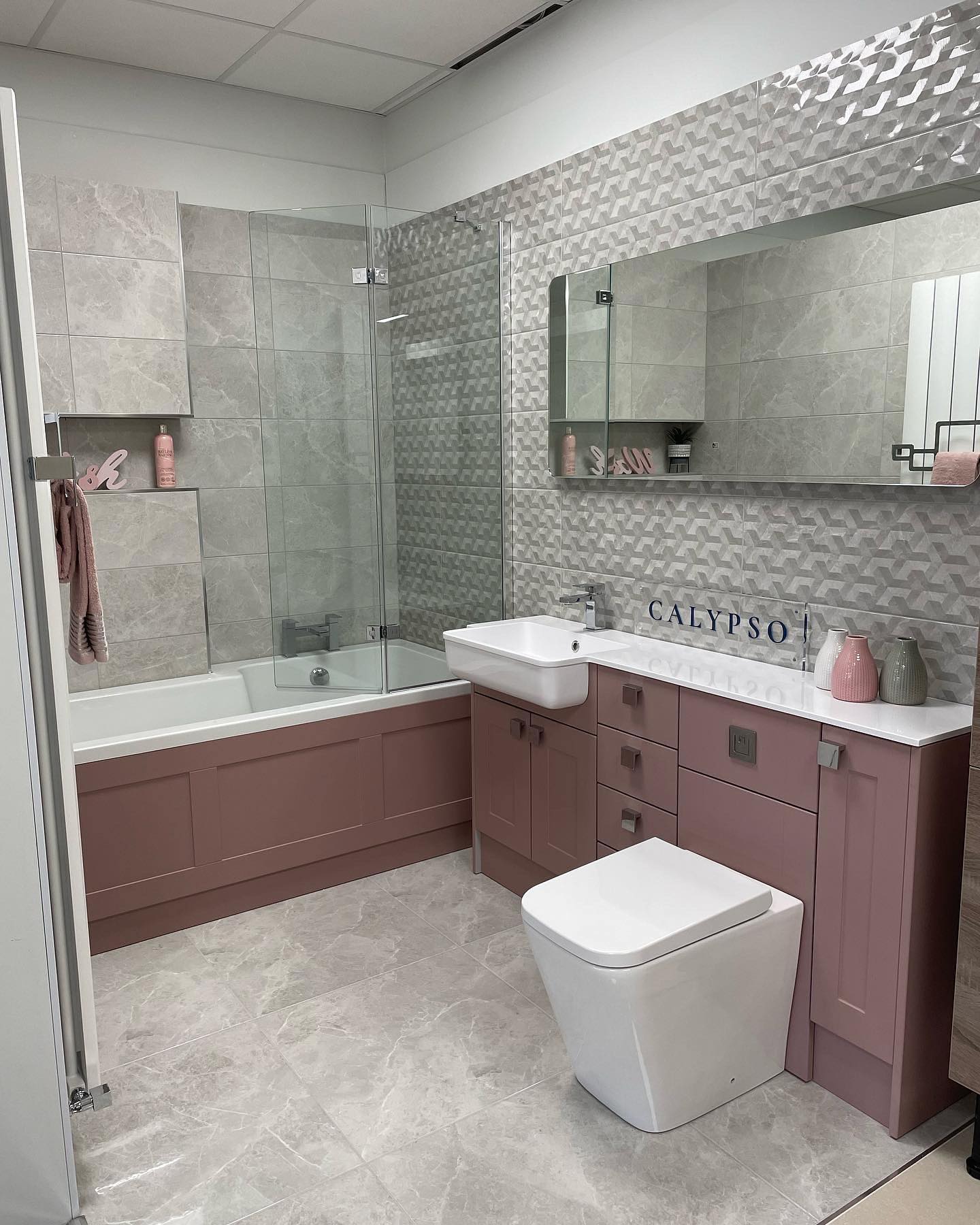 Showroom display with dusky pink fitted furniture, a bath with glass screen and grey tiles